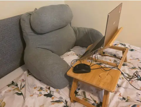 Gillian Cooley A large support cushion leans against a grey headboard and a laptop sits on a small desk on the bed. 