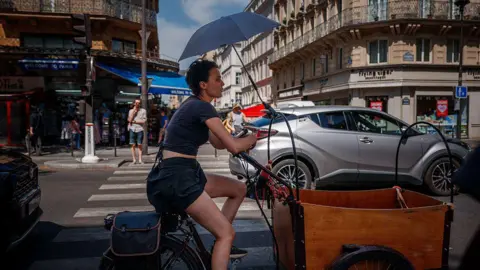 Dimitar DILKOFF / AFP via Getty Images A woman protects herself from the sun under an umbrella as she rides a bicycle in Paris on June 25, 2026, as France experiences a heatwave.