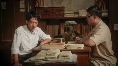 Golden Village A man sits across another man at a desk. One of them is writing on a piece of paper.