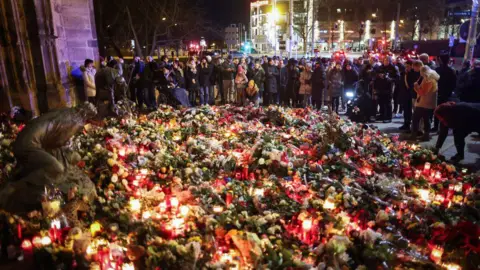 AFP via Getty Images A large crowd gather at a red-and-white cordon at night, holding candles for a vigil. The front row mostly comprises women, all of whom wear large winter coats. It is night time. They are standing in front of a sea of flowers - many bouquets heaped on top of each other, stretching for several metres in every direction.