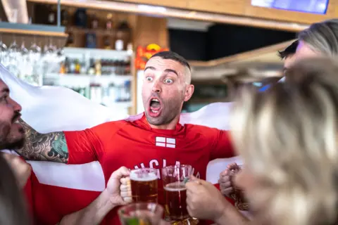 Getty Images A young man, wearing an England T-shirt, is celebrating in the pub with friends. He is lifting an England flag behind him, worn like a cape. His friends are chinking pints of beer. 