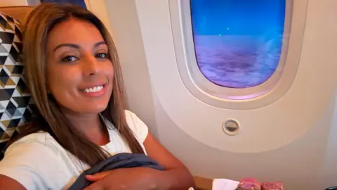 Karis Heemskerk Karis smiles while sitting in an aircraft seat, a dusky blue sky with a thin layer of clouds outside the window.