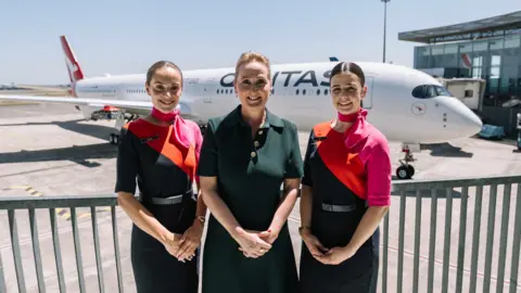 Qantas Three woman stand with their hands clasped in front of them in front of a large aircraft with 'Qantas' painted down the fuselage.