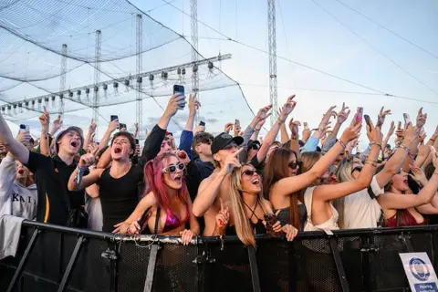 Redferns/Getty Images Festivalgoers attend Leeds Festival at Bramham Park. They are shown with their hands in the air cheering and smiling, at the barriers in a performance.