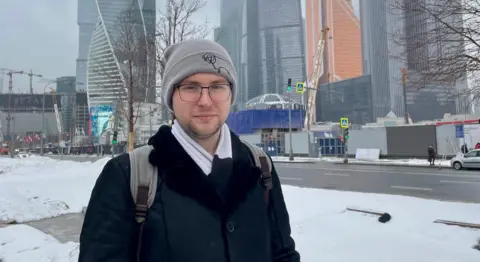 Ben/ BBC Person wearing a grey beanie, glasses, a black coat and a white scarf, carrying a grey backpack with visible shoulder straps in the foreground. Snow covers the pavement and roadside. A wide street with traffic lights and pedestrian crossing signs runs behind. In the background, modern high-rise buildings with glass façades and construction cranes are visible. Bare trees line the street, and temporary site structures and barriers are present near the road.