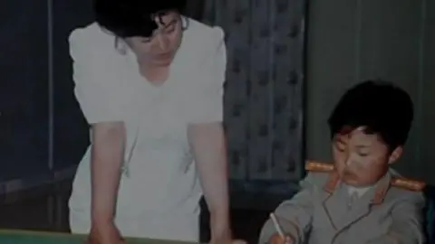Cheong Seong-chang Ko Yong Hui, in a white dress, stands on the left. A young Kim Jong Un, in a miitary uniform, sits and writes with a pen. 