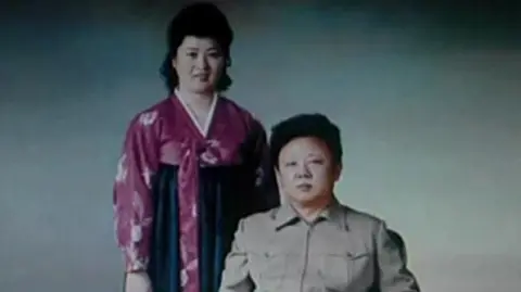 Cheong Seong-chang Ko Yong Hui, in a purple and green Korean dress stands on the left. Kim Jong Il sits besides her 