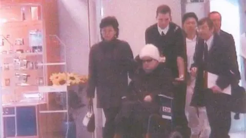 Yoji Gomi Ko Yong Hui, in sunglasses and a white beanie, sits in a wheelchair pushed by a man. Several people are around her at a hospital in France