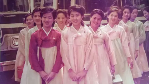 Yoji Gomi About a dozen of young women in traditional Korean dresses stand in front of vehicles. Ko Yong Hui stands at the front, on the left 