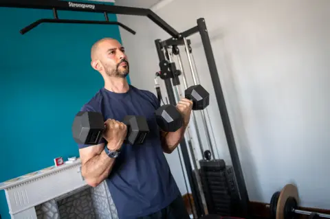 BBC/Emma Lynch Marios in a dark blue t-shit and shorts doing bicep curls with two dumbbells 
