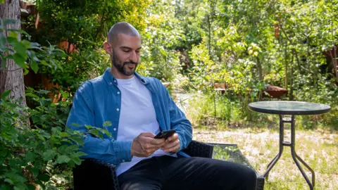 BBC/Emma Lynch A man with a dark moustache and beard in a blue shirt, white t-shirt and back trousers, sitting in a garden, looking at his mobile phone. 