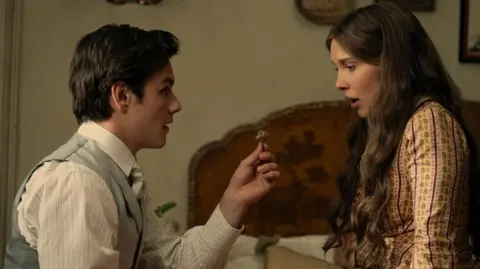 Netflix Louis Partridge and Millie Bobby Brown in Enola Holmes. He is holding a ring and she is sitting on the bed looking surprised