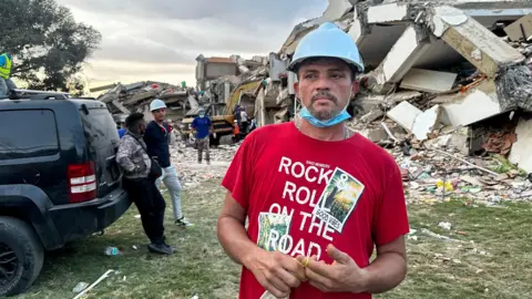 Ronnie Navarro wearing a red flannel top and a blue helmet with the pile of rubble in the background.