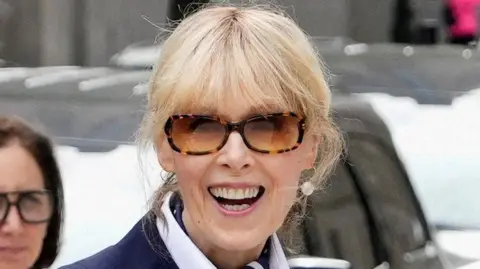 REUTERS/Adam Gray Writer E. Jean Carroll is seen outside looking straight at the camera and smiling. She has blonde hair pulled back and wears large pearl earrings, subglassess and a white shirt with a blue blazer. 