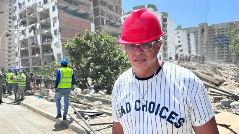 A man in a red hard hat and sunglasses stands on a street in front of the rubble of a collapsed building, with officials in high-visibility vests nearby.