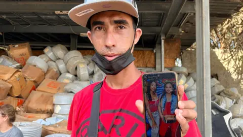 A man in a baseball cap and red t-shirt displays a photo of his wife and daughter on a mobile phone he holds aloft in his left hand.