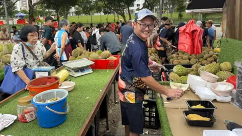 Kelly Ng/ BBC A man smiling at the camera as he cuts durian