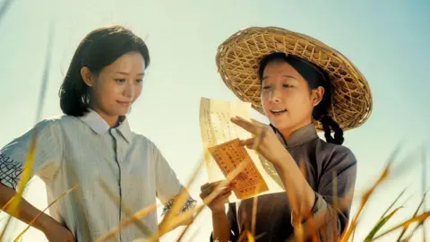 Golden Village Two women standing in a field, one is reading a letter out to the other.