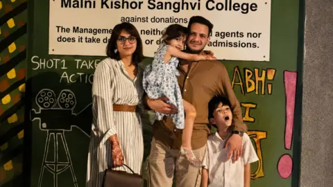 Imtiyaz Ali From left: Mariam, dressed in a striped dresss with glasses on, stands next to Javed, who is wearing a brown shirt and khaki pants, and holding their daughter dressed in frock, along with their son, who leans on to his father with his mouth open in a comical expression. 