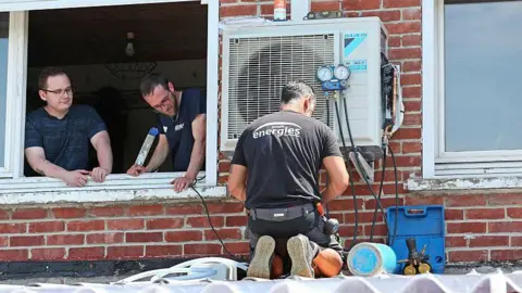 AFP via Getty Images A man in black clothes with his back to the camera installs an air conditioning unit at a home in Mericourt, northern France on 19 June 19, 2026. From a window, two men look at him working 