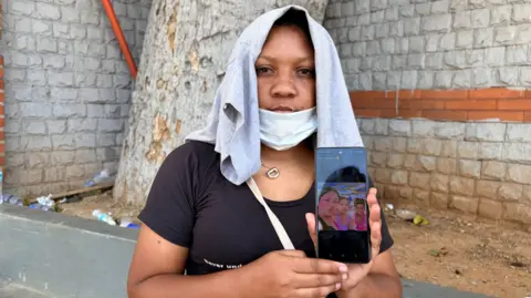 BBC A woman with a grey t-shirt covering her head holds up a mobile phone to display a photo of her with her two teenage daughters.