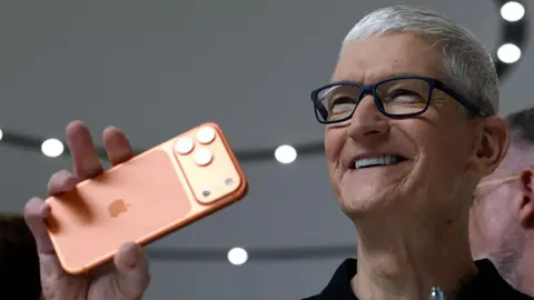 Getty Images Apple CEO Tim Cook smiles as he holds up an orange iPhone 17 Pro during an event at the company's headquarters 