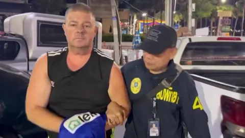 Reuters Simon Peter Carman, a man with short light brown hair wearing a sleeveless black too, is escorted by a Thai police officer.
