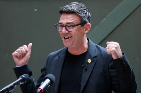 Reuters Andy Burnham, wearing a black jacket and t-shirt, stands at a podium.