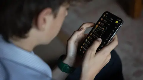 Getty Images A photo illustration showing a teenage boy looking at his phone as he scrolls through the Snapchat app. He is wearing a watch on his left wrist, and has short black hair. 