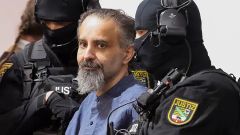 Reuters Taleb al-Abdulmohsen, a man with greying brown hair and a greying beard, looks on while being escorted by armed security officers. He wears a light blue buttoned shirt. The security officials wear all-black uniforms, including face coverings with only the eyes visible.