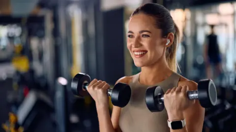 Getty Images A woman in a gym, who is holding two dumbells and smiling towards the camera. She has a blonde ponytail and is wearing gym clothes.