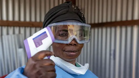 Getty Images A healthworker, wearing goggles and a mask, pulled down from her mouth, holds up a thermometer