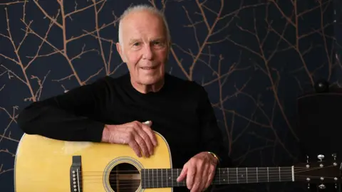 BBC Allan Taylor seated and looking at the camera and smiling with a guitar o his lap and both arms resting on it.