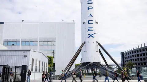 Getty Images A row of people walk past a large white rocket with blue "space x" logo written on it.