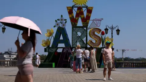 Reuters A woman walks under an umbrella past a "Warsaw" sign as a heatwave sweeps Poland, in Warsaw, Poland, 
