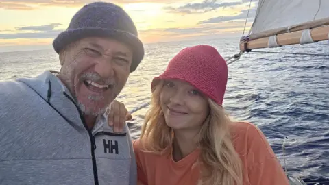 Rob Donald Man with moustache and trimmed beard is wearing a fleece and knitted wool hat. Sitting next to him is a girl with her hair flying in the wind. She has a red wool hat on. She is smiling and they are both on a yacht. 