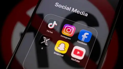 Getty Images TikTok, Instagram, Facebook, X, Snapchat and YouTube apps displayed on an iPhone screen under a "social media" tab, with an icon used to show under-16s banned reflected across it.