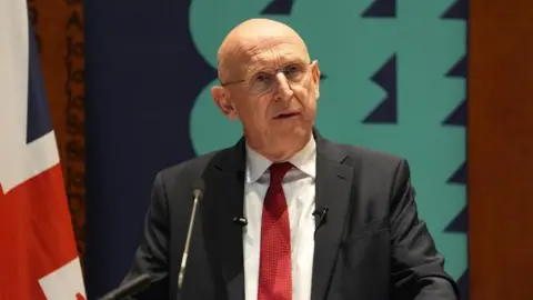 PA Media John Healey wearing a dark suit and red tie standing before a microphone