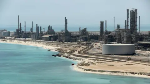 Reuters A file photo of Aramco's Ras Tanura oil refinery and oil terminal, which lies along a stretch of coast. The site comprises a network of several tall chimney-shaped structures and smaller buildings. It is daytime and the water along the coastline is a clear blue.
