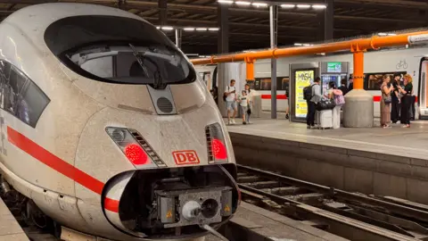 Reuters A DB Deutsche Bahn passenger train is seen at a standstill as passengers with suitcases wait on the platform to the right.