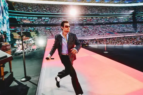 Anthony Pham Harry Styles strides onto the stage at Wembley Stadium, wearing a black and white pinstripe jacket and a wide-collared blue shirt. Spotlights illuminate the audience, and the singer is wearing aviator sunglasses.