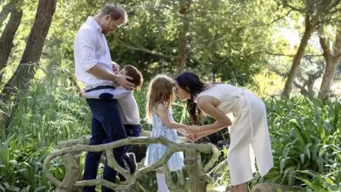Instagram/Meghan, Duchess of Sussex Archie is hugging his father, Prince Harry, the Duke of Sussex, while Meghan is bent to eye-level and holding hands with Lilibet, as they all stand on what appears to be carved mini wooden bridge over a brook among trees.