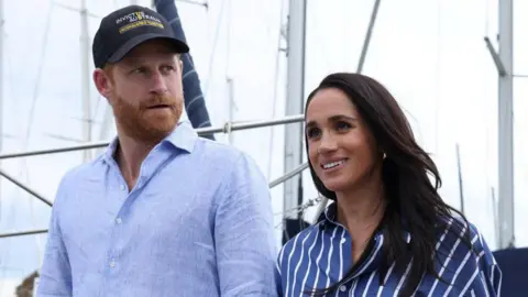 Reuters Prince Harry and Meghan walking next to each other, both in blue shirts as sails of boats are visible behind them in Sydney Harbour. Photo taken April 17 2026.