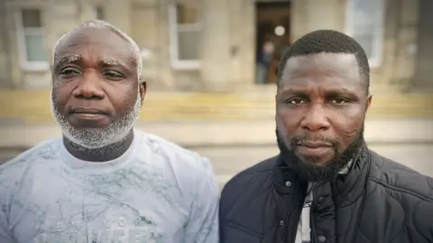 BBC Two men shown side-by-side in a split portrait, both facing the camera outdoors in front of a blurred building. The man on the left has a short haircut with a grey beard and is wearing a light-coloured patterned shirt, while the man on the right has short hair, a trimmed beard, and is wearing a dark quilted jacket over a shirt.