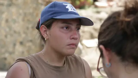 BBC Andreina Valerio, in a blue baseball cap, stands in front of rubble