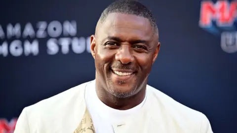 Getty Images Luther Idris in a cream jacket and white T-shirt