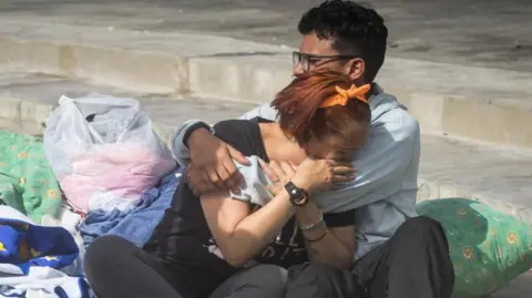 EPA Two people cry into each others' arms surrounded by pillows, blankets and carrier bags of possessions on concrete stairs outside