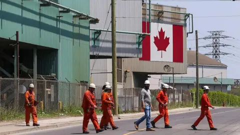 AFP via Getty Images A Canadian national flag is seen in the background as seven workers dressed in orange work suits and white hard hats cross the street in front of ArcelorMittal Dofasco's steel manufacturing buildings in Hamilton, Ontario. 