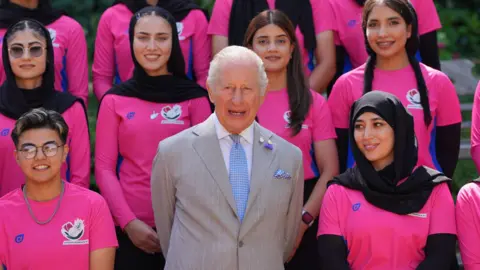 PA Media King Charles wearing a pale grey suit with white shirt and blue tie, with members of the Afghan women's cricket team - wearing hot pink T-shirts - on either side and behind him. All are looking ahead and smiling 