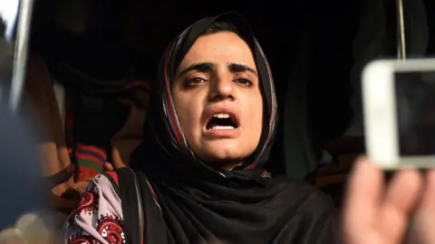EPA Mahrang Baloch, a woman, is pictured mid-speech with a serious expression. A hand holding a mobile phone aloft, as if to record her speech, is pictured to the right of the frame.